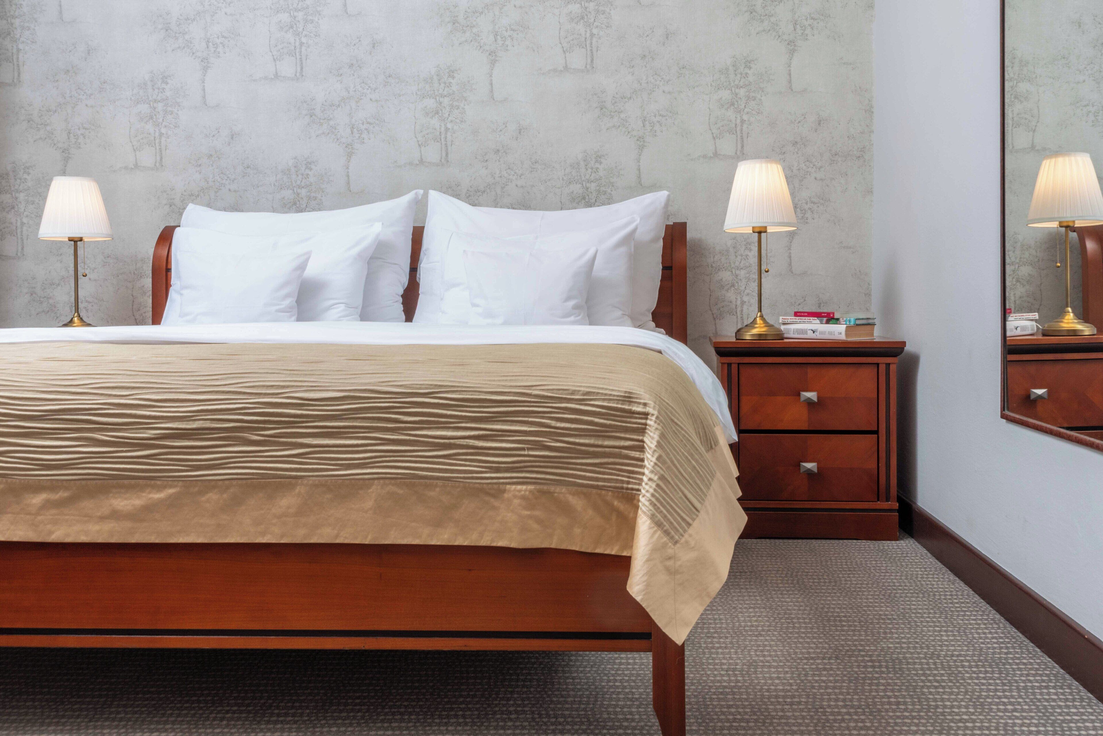 Una cama bien hecha con sábanas blancas, dos lámparas de noche, una mesita de madera con espejo y una pared decorada.
