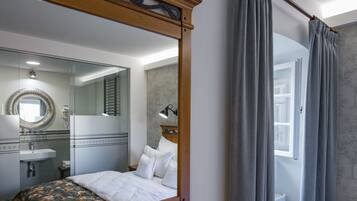 Un dormitorio con un espejo grande, una cama con sábanas blancas, una mesita de noche y una ventana con cortinas.