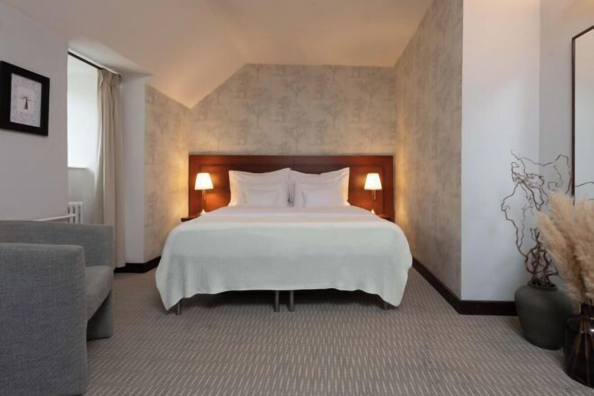 A hotel room with a bed, two bedside lamps, a grey armchair, a large window, and a wall-mounted artwork.