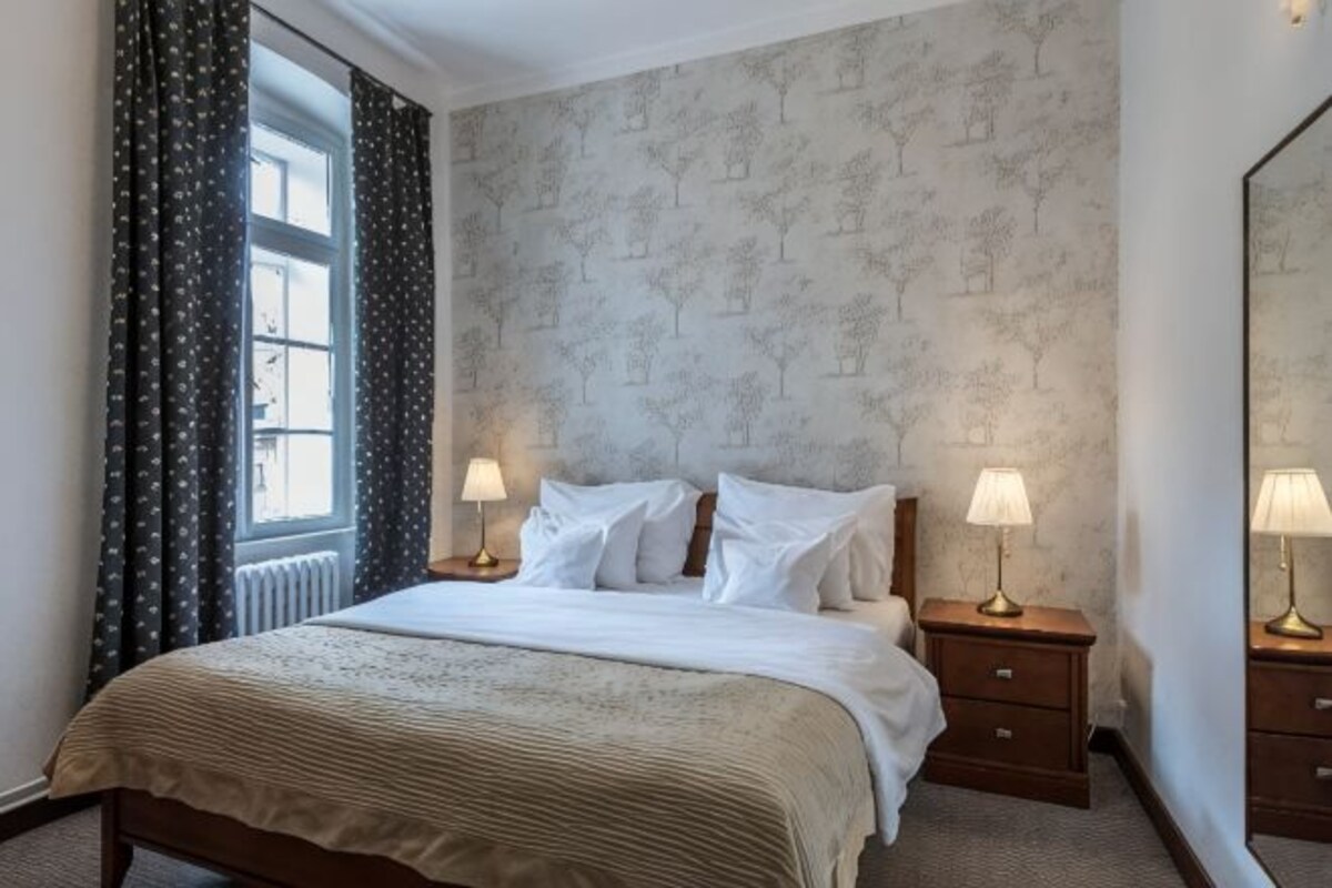 Habitación de hotel con una cama grande, dos mesitas de noche con lámparas, una ventana con cortinas y un papel tapiz estampado.