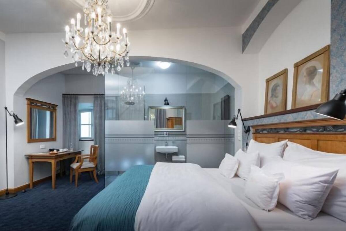 A hotel room with a large bed, a chandelier, a desk, and a mirror.