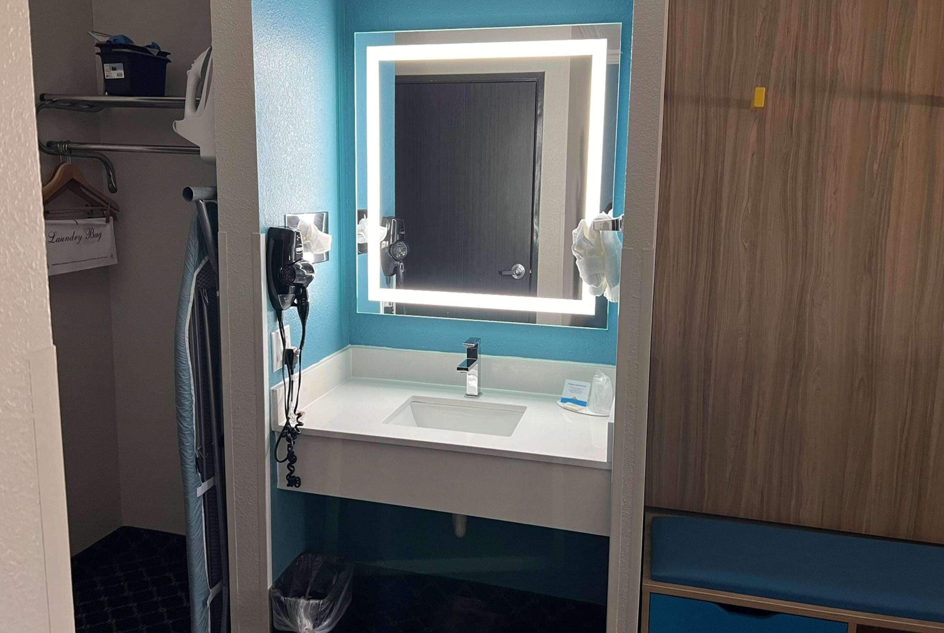 A bathroom with a white sink, a mirror with a light, a towel rack, and a trash bin.