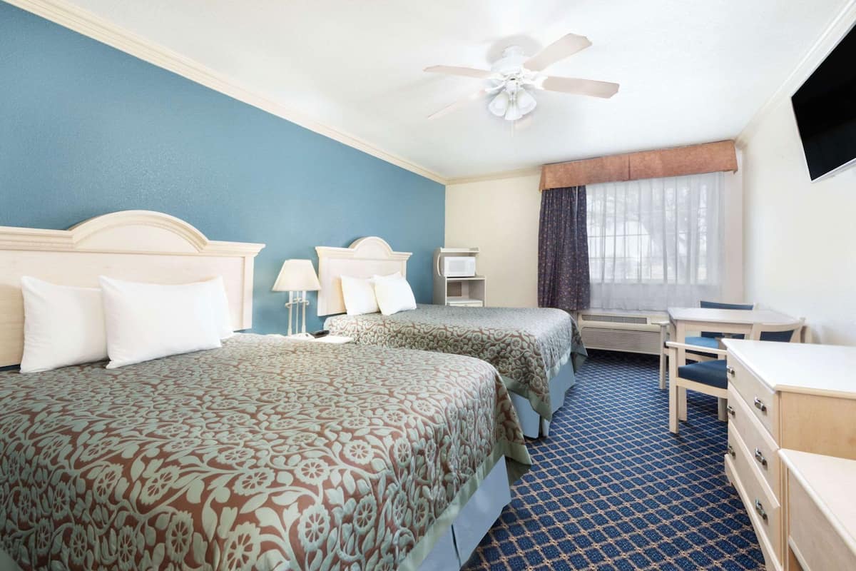 Habitación de hotel con dos camas, un escritorio, una silla, un ventilador de techo y una ventana con cortinas.