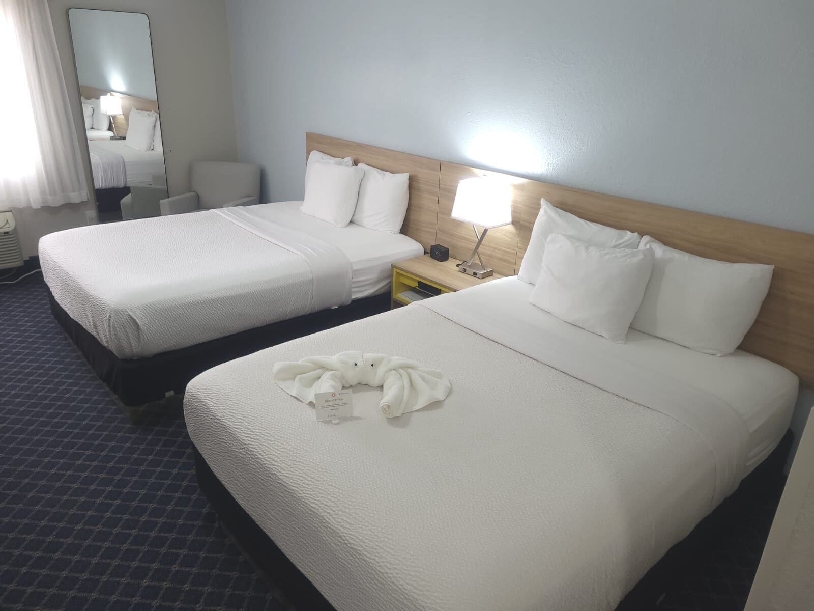 A hotel room with two beds, a nightstand, a mirror, and a window with curtains.