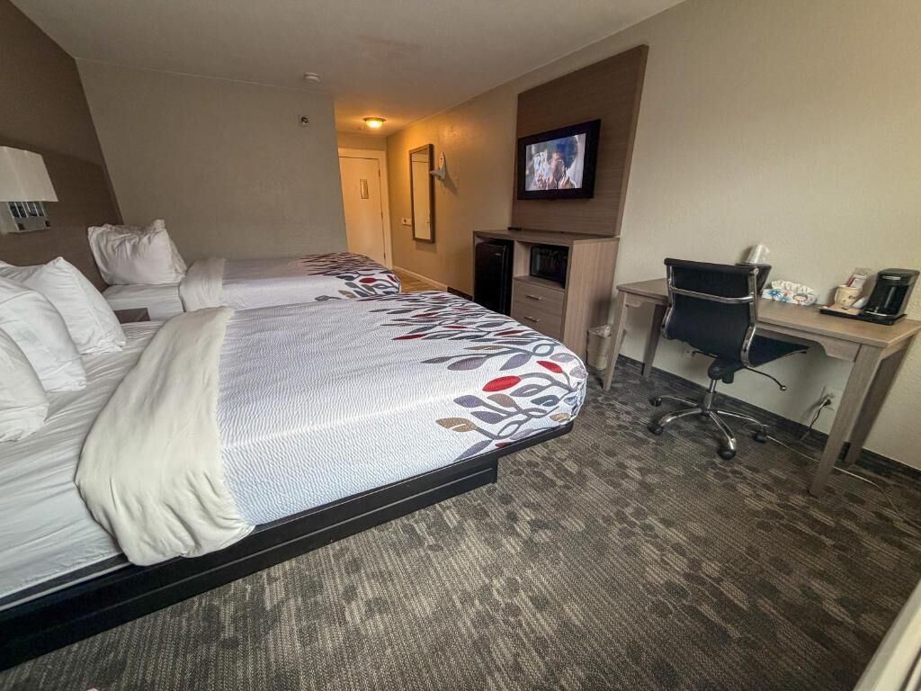 A hotel room with two beds, a desk, a chair, a television, and a small kitchenette.