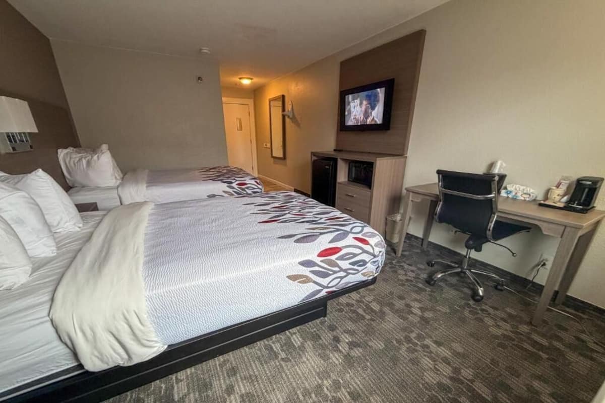 A hotel room with two beds, a desk, a chair, a television, and a small kitchenette.