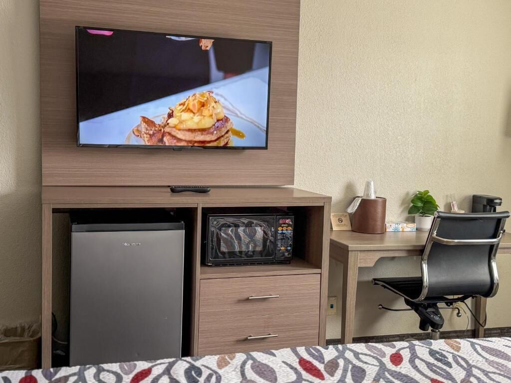 A hotel room with a TV mounted on a wooden cabinet, a microwave, a refrigerator, a desk with a chair, and a bed with a patterned bedspread.