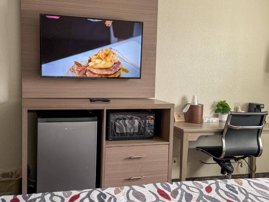 Et hotelværelse med et fjernsyn monteret på et træskab, en mikrobølgeovn, et køleskab, et skrivebord med stol og en seng med et mønstret sengetæppe.