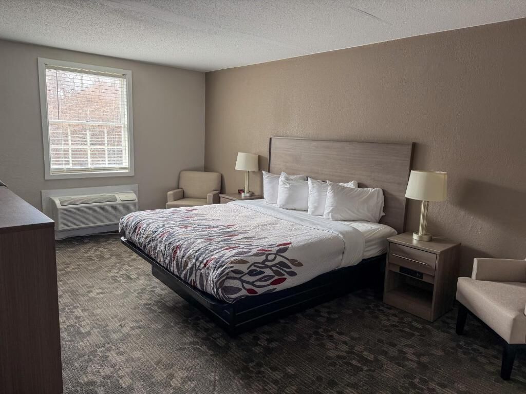 A hotel room with a bed, two bedside tables, a chair, a window with blinds, and an air conditioning unit.