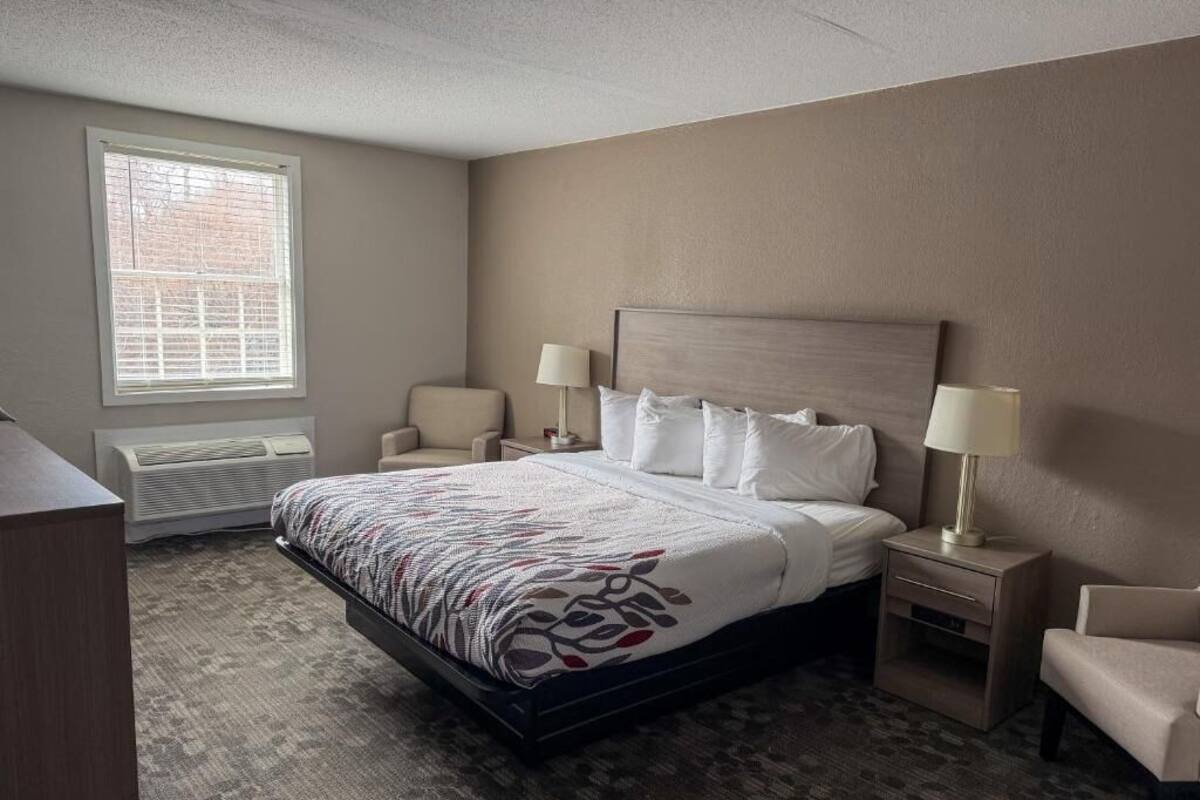 Una habitación de hotel con cama, dos mesitas de noche, una silla, ventana con persianas y aire acondicionado.