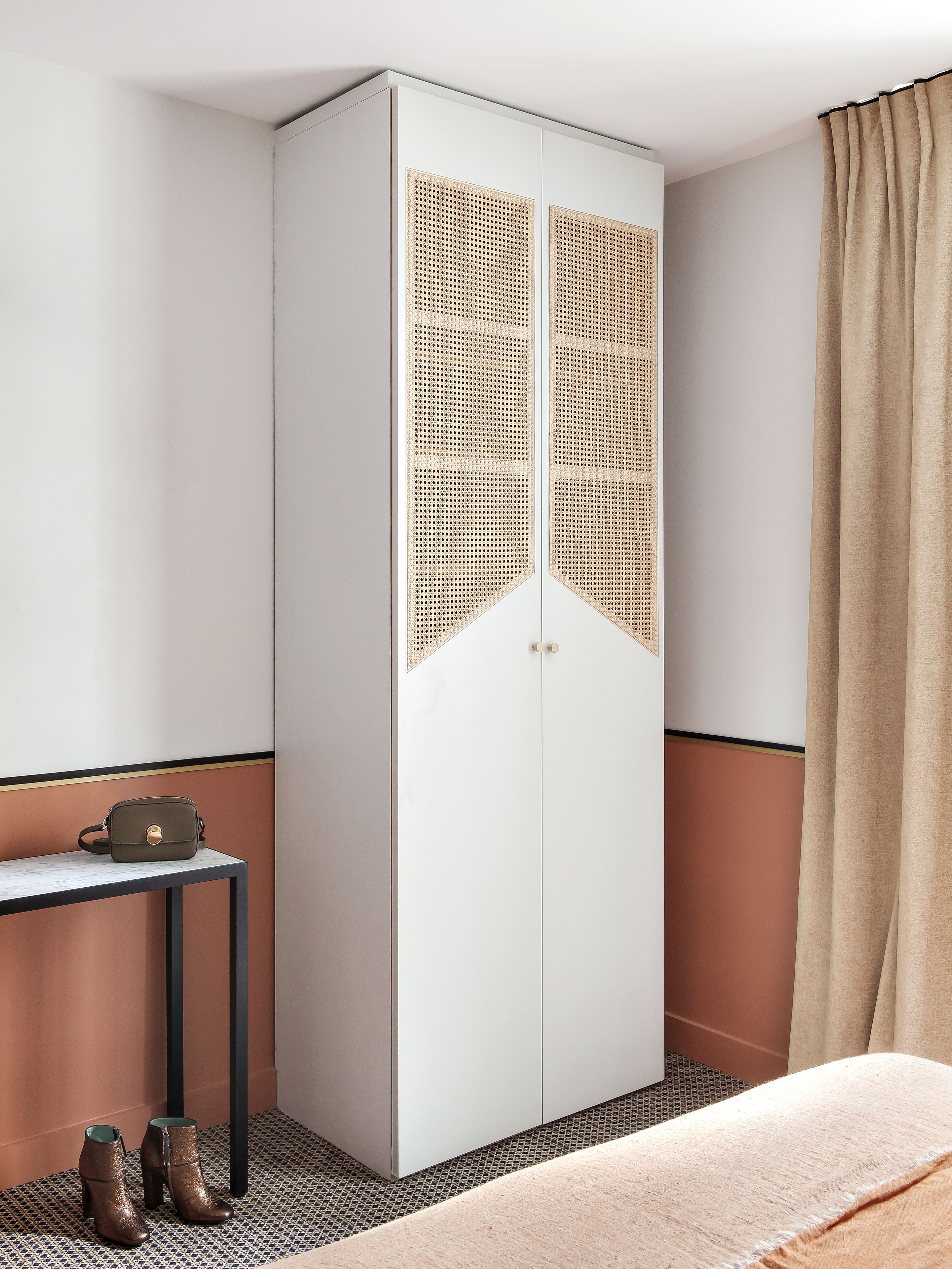 Una habitación con un armario blanco con diseño de panel tejido, una mesita con un bolso y botas, y una cama con cubierta color beige.