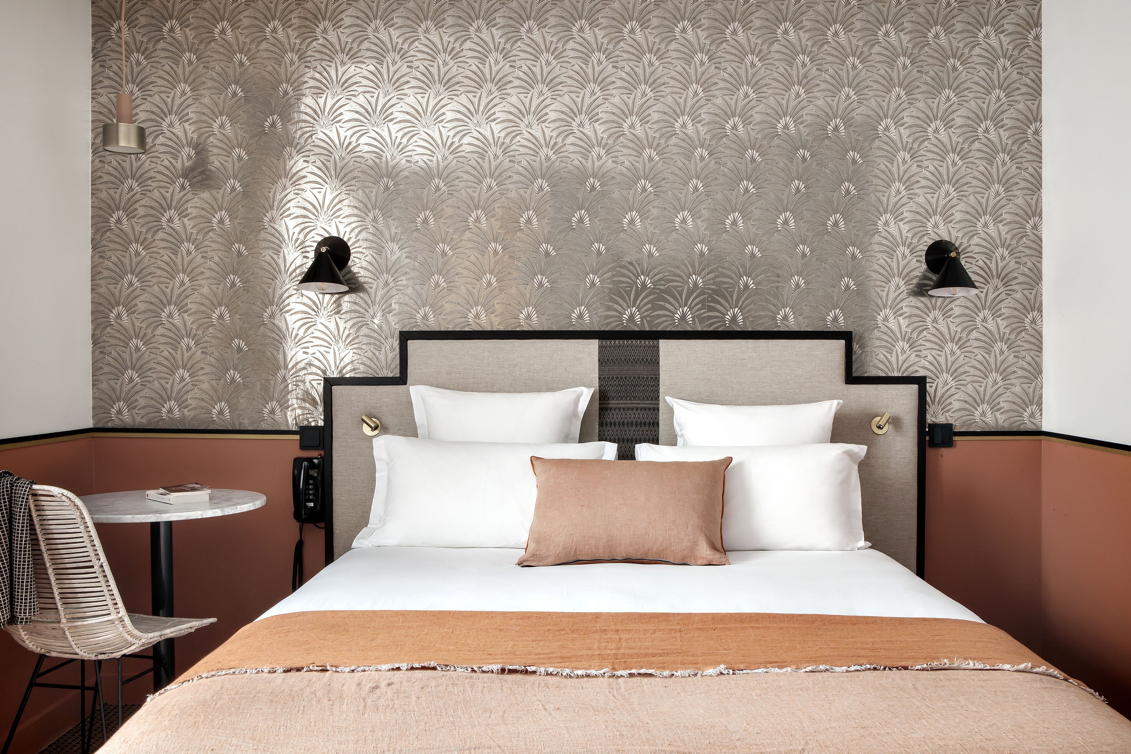 Habitación de hotel con cama, dos lámparas de noche, una mesita con silla y un papel tapiz estampado.