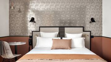 Habitación de hotel con cama, dos lámparas de noche, una mesita con silla y un papel tapiz estampado.