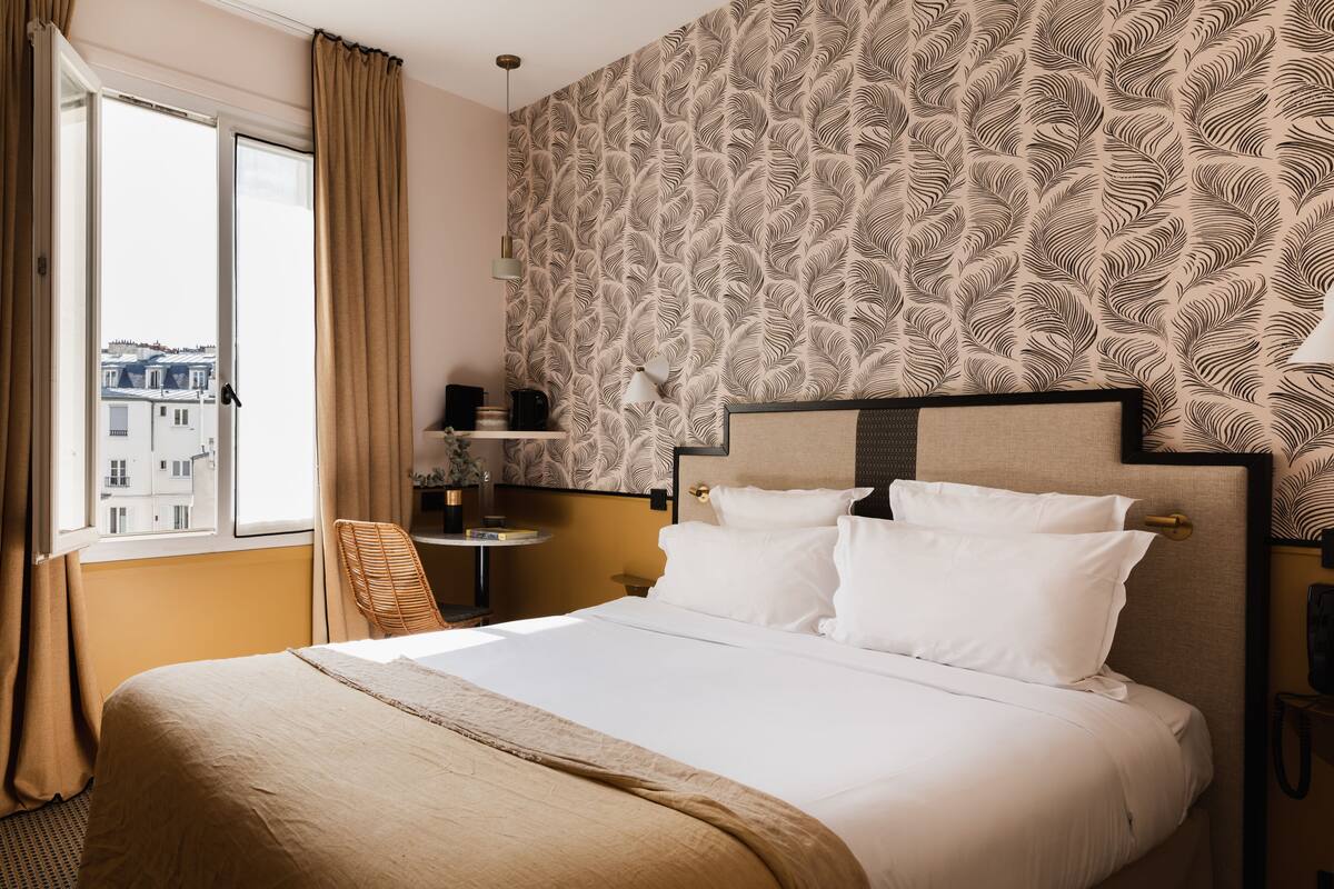 Quarto de hotel com uma cama grande, janela com cortinas, papel de parede estampado e uma mesa pequena com abajur.