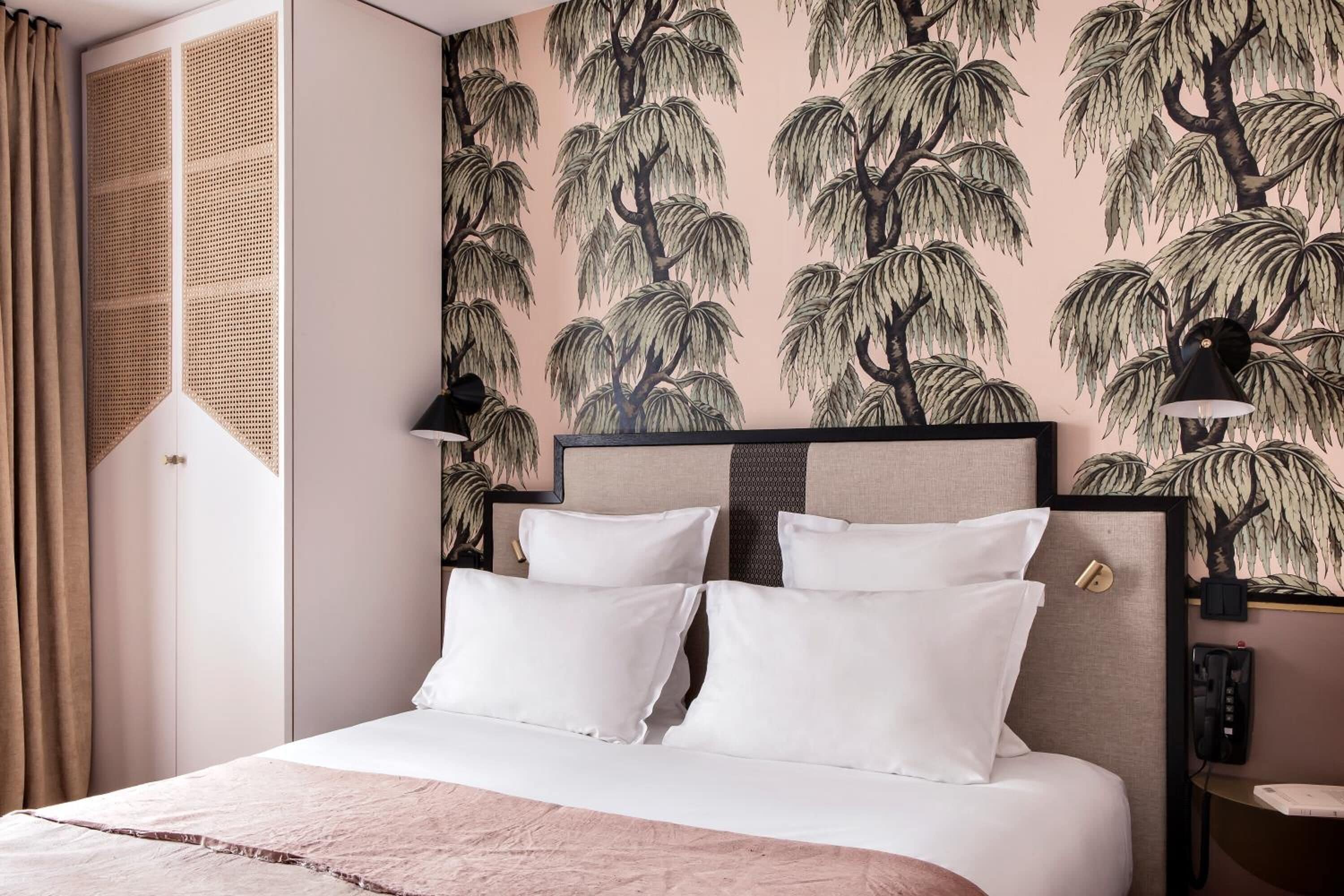A neatly made bed with white linens and pillows, a headboard with a geometric pattern, and a wall with a tree and palm leaf wallpaper.