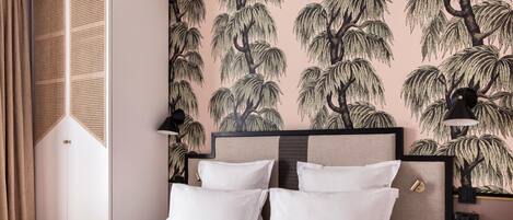 Una cama bien hecha con sábanas y almohadas blancas, cabecera con diseño geométrico, y pared con papel tapiz de hojas de árbol y palmera.