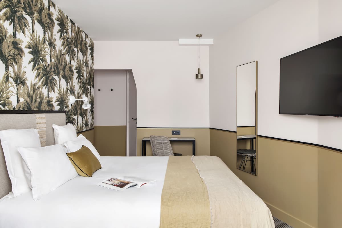 Quarto de hotel com cama, escrivaninha, poltrona, televisão e parede com papel de parede de palmeira.