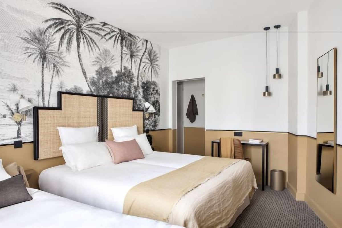 A hotel room with a large bed, a bedside table, a chair, a small desk, a trash bin, a wall-mounted mirror, and a wall art depicting palm trees.