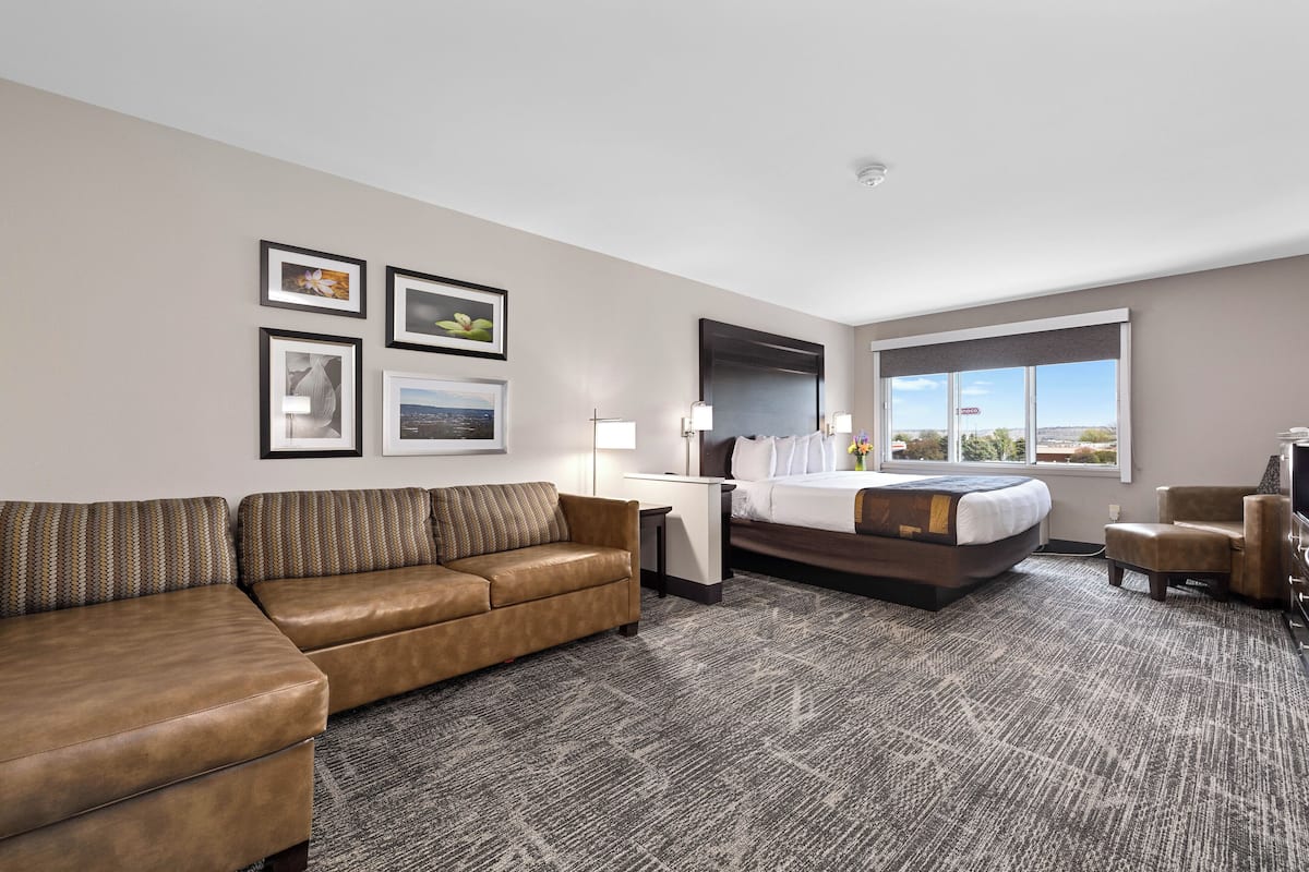 A hotel room with a brown leather sofa, a bed with white linens, a desk with a lamp, and framed pictures on the wall.