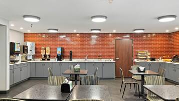 A modern kitchen area with grey cabinets, a brick backsplash, and a central island with a sink. There are multiple tables and chairs, and a coffee machine is visible.
