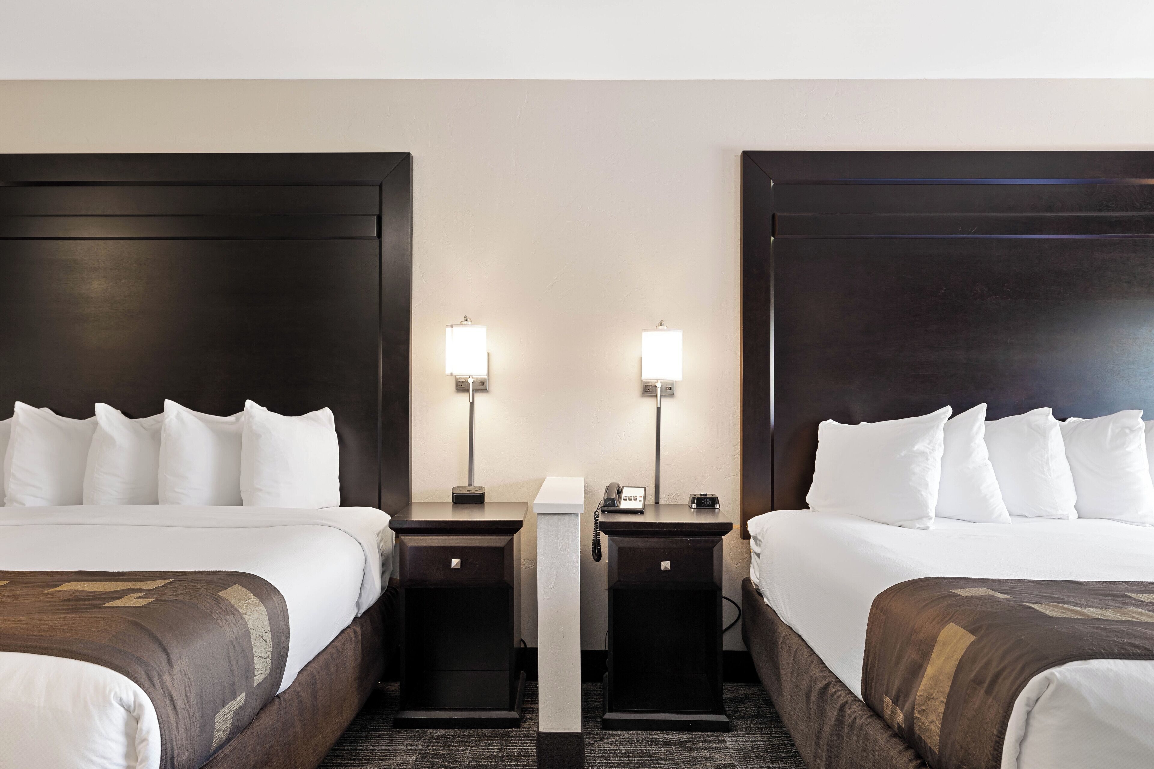 A hotel room with two beds, each with white linens and dark headboards. There are bedside tables with lamps and a phone on one side.