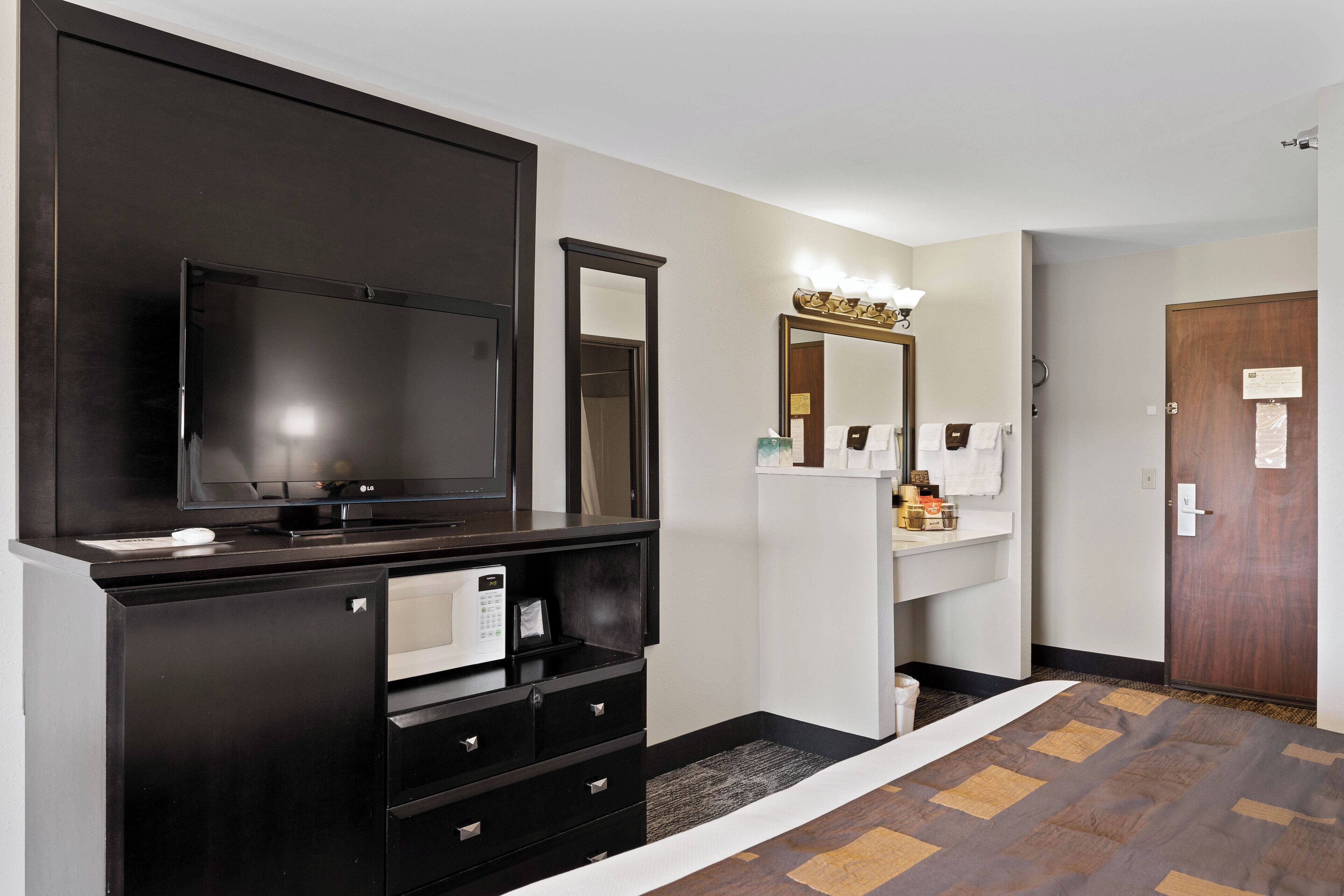Habitación de hotel con un televisor de pantalla plana grande, una cómoda de madera oscura con espejo, un lavabo blanco y una puerta con panel de madera.