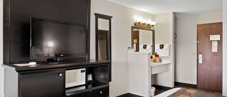 Habitación de hotel con un televisor de pantalla plana grande, una cómoda de madera oscura con espejo, un lavabo blanco y una puerta con panel de madera.