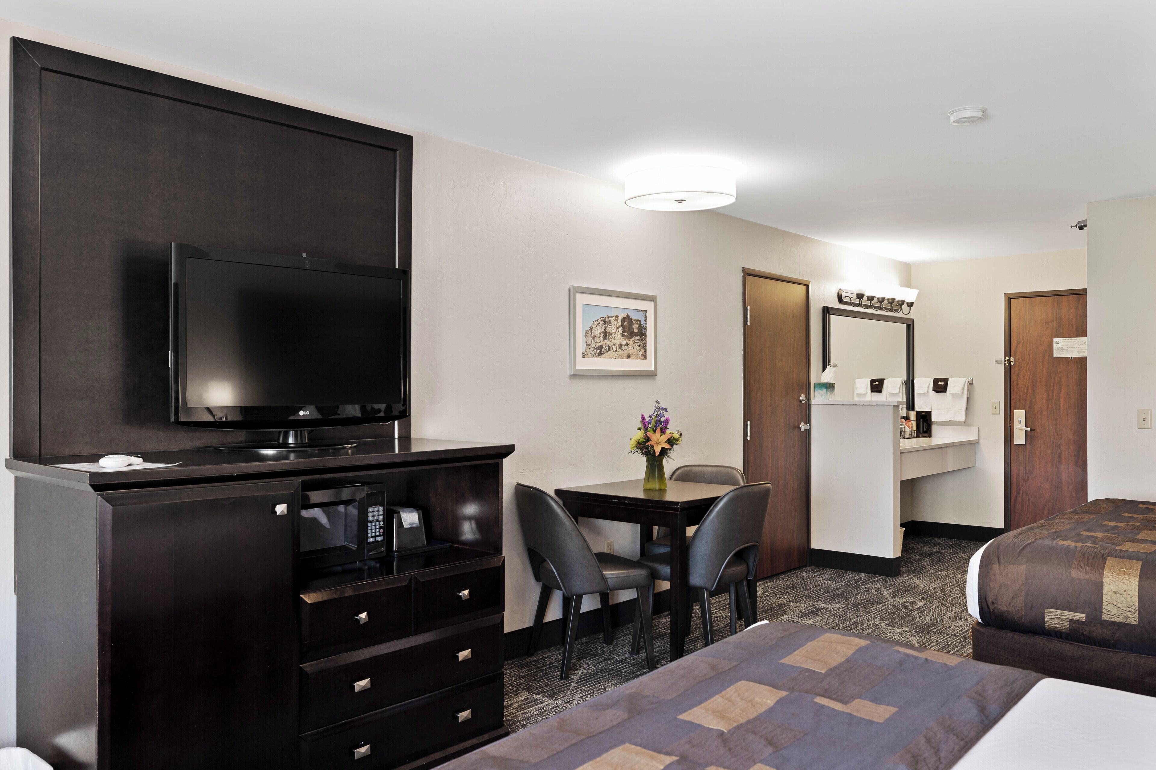 Une chambre d’hôtel équipée d’une télévision à écran plat, d’un bureau avec deux chaises, d’une commode, d’un lit et d’une salle de bain avec un miroir et un lavabo.