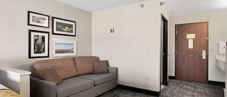 A hotel room with a grey sofa, a bed with a patterned bedspread, a carpeted floor, and a wooden door.
