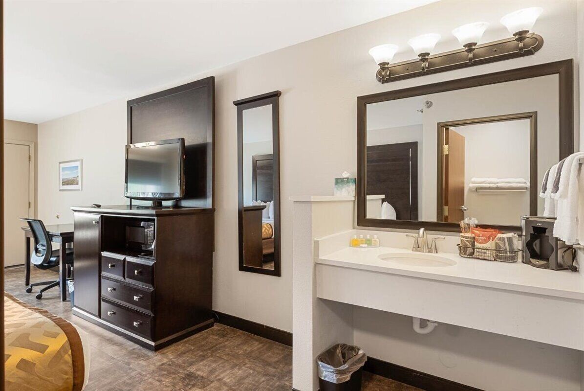 A hotel room with a dark wood dresser, a flat-screen TV, a white vanity with a mirror, and a small trash can.