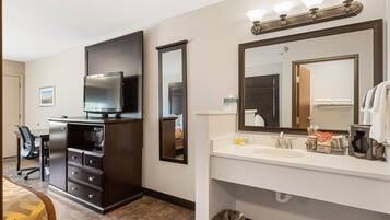 A hotel room with a dark wood dresser, a flat-screen TV, a white vanity with a mirror, and a small trash can.