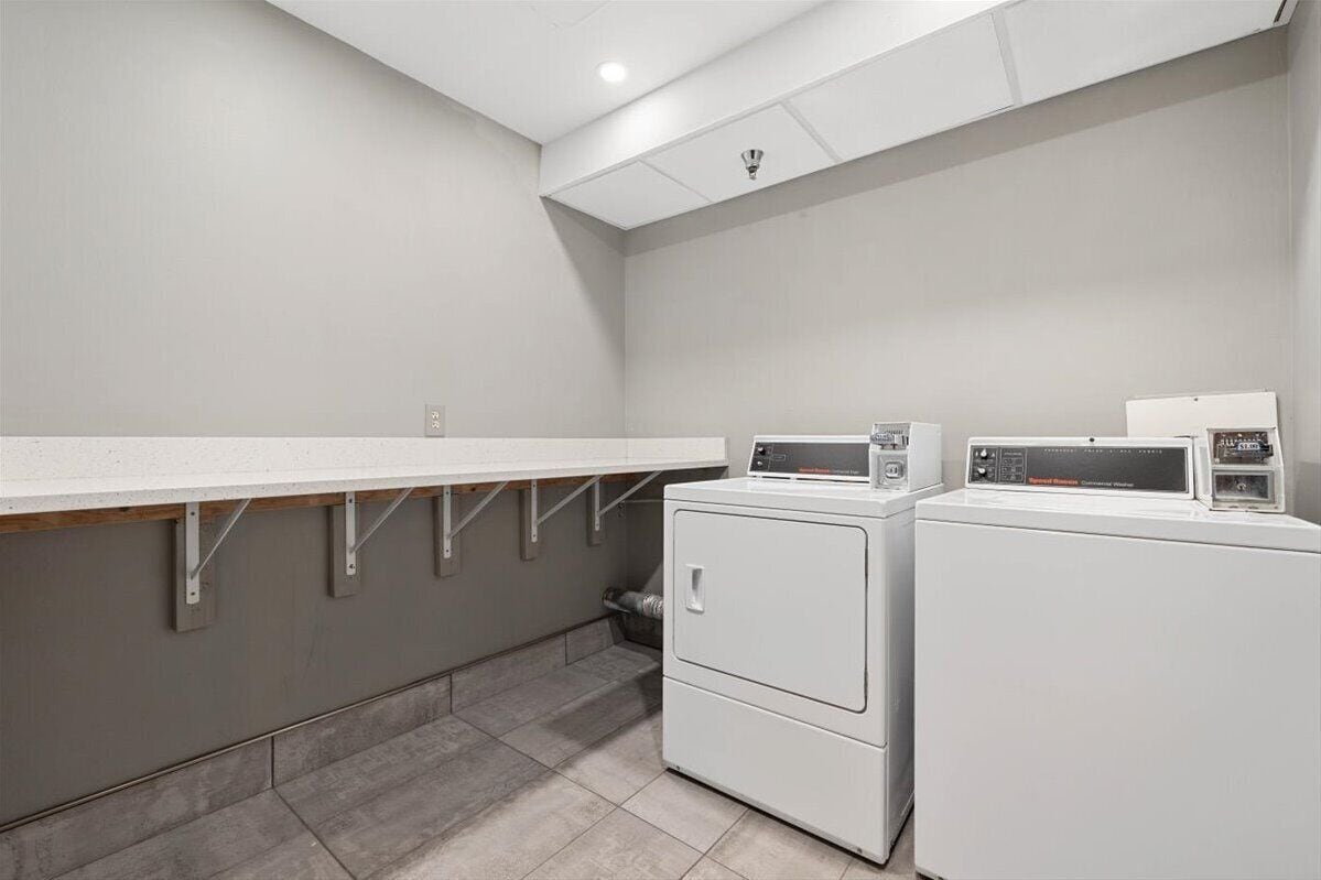 A laundry room with two washing machines and a drying machine, a long counter, and a tiled floor.