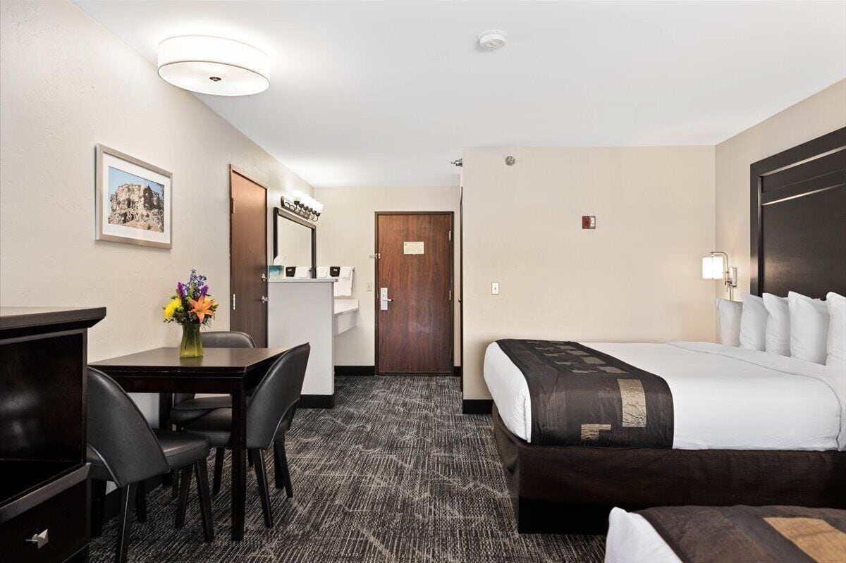 A hotel room with a bed, a desk with chairs, a small table with a vase of flowers, and a door.