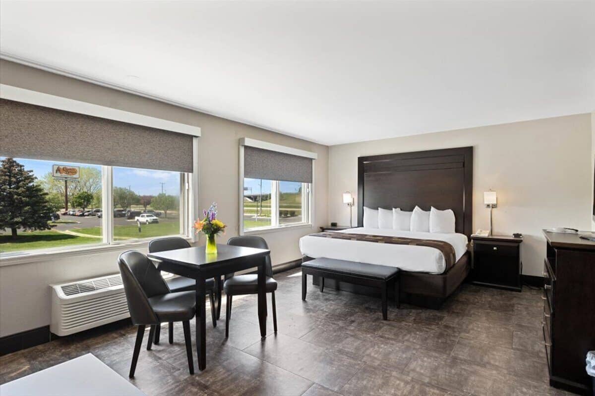 A hotel room with a large bed, a desk with chairs, a bench, a nightstand, and a view of the parking lot through the windows.