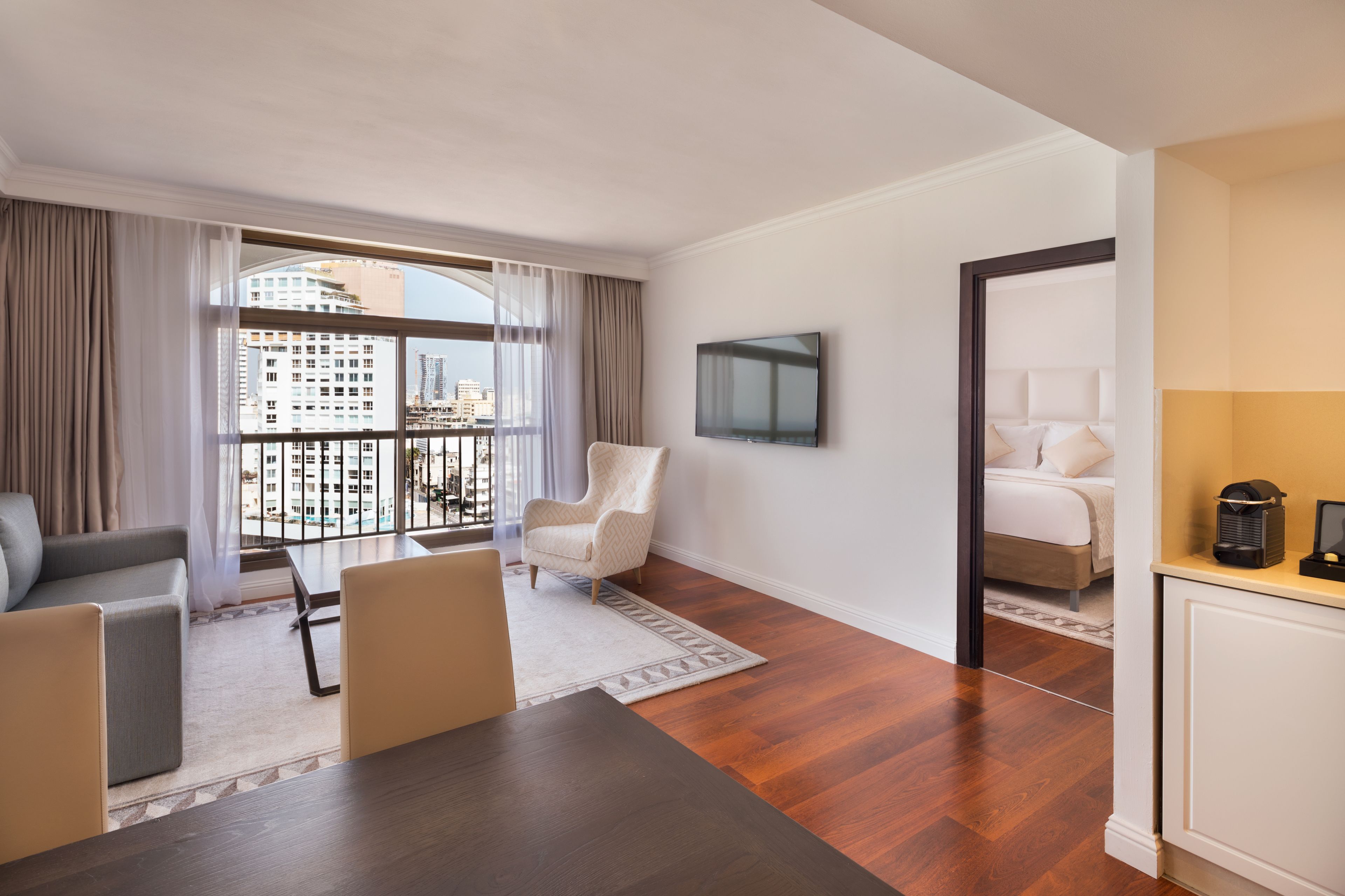 A modern hotel room with a large window, a flat-screen TV, a bed, a dining table, and a seating area with a sofa and chairs.