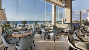 A modern lounge area with large windows overlooking a beach and ocean, furnished with round tables and chairs. - Orchid Tel Aviv (Tel Aviv)
