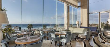 A modern lounge area with large windows overlooking a beach and ocean, furnished with round tables and chairs.