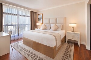 Habitación de hotel con una cama grande, un escritorio, una silla, una mesita de noche con lámpara y vistas a la ciudad a través de cortinas translúcidas.