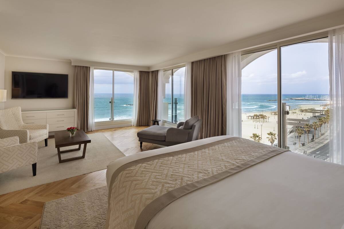 A spacious hotel room with a large bed, a sofa, a TV, and a view of the beach and ocean.