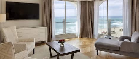 A spacious living room with a large TV, a sofa, a coffee table with a vase, and a view of the ocean.