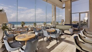 A modern outdoor seating area with round tables and chairs, overlooking a beach and ocean. - Orchid Tel Aviv (Tel Aviv)
