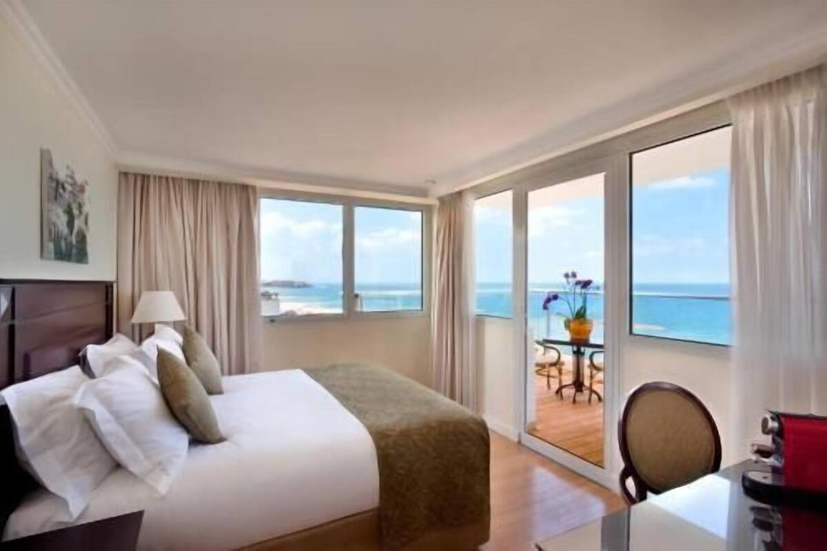 A hotel room with a bed, a desk, a chair, a table with a vase, and a view of the sea.
