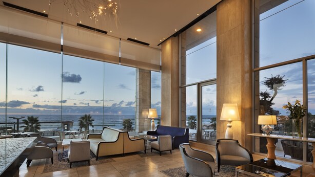 A spacious lounge area with a large window offering a view of the ocean and palm trees. There are several seating options including sofas and chairs, and a small table with a lamp. - Orchid Tel Aviv (Tel Aviv)