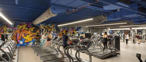 Uma academia com esteiras, equipamentos de exercício e um mural colorido na parede.