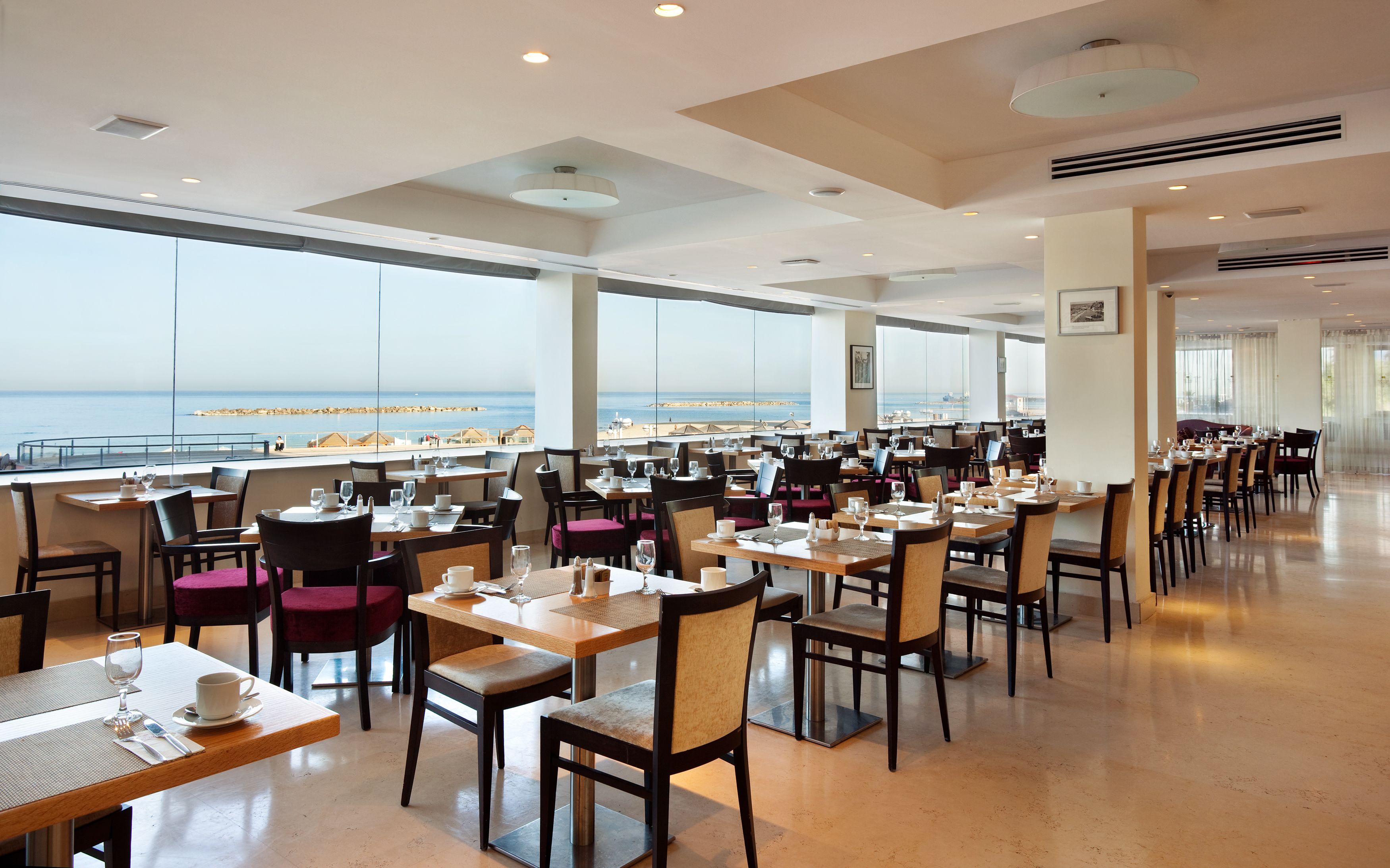 A spacious dining area with tables and chairs, large windows offering a view of the beach and ocean, and a modern ceiling design with recessed lighting.