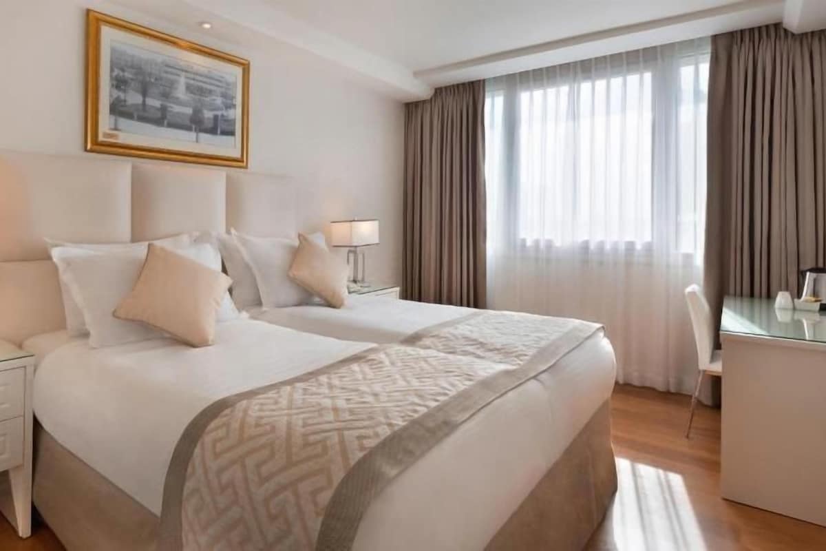 Una habitación de hotel con dos camas, un escritorio, una silla y un ventanal con cortinas.