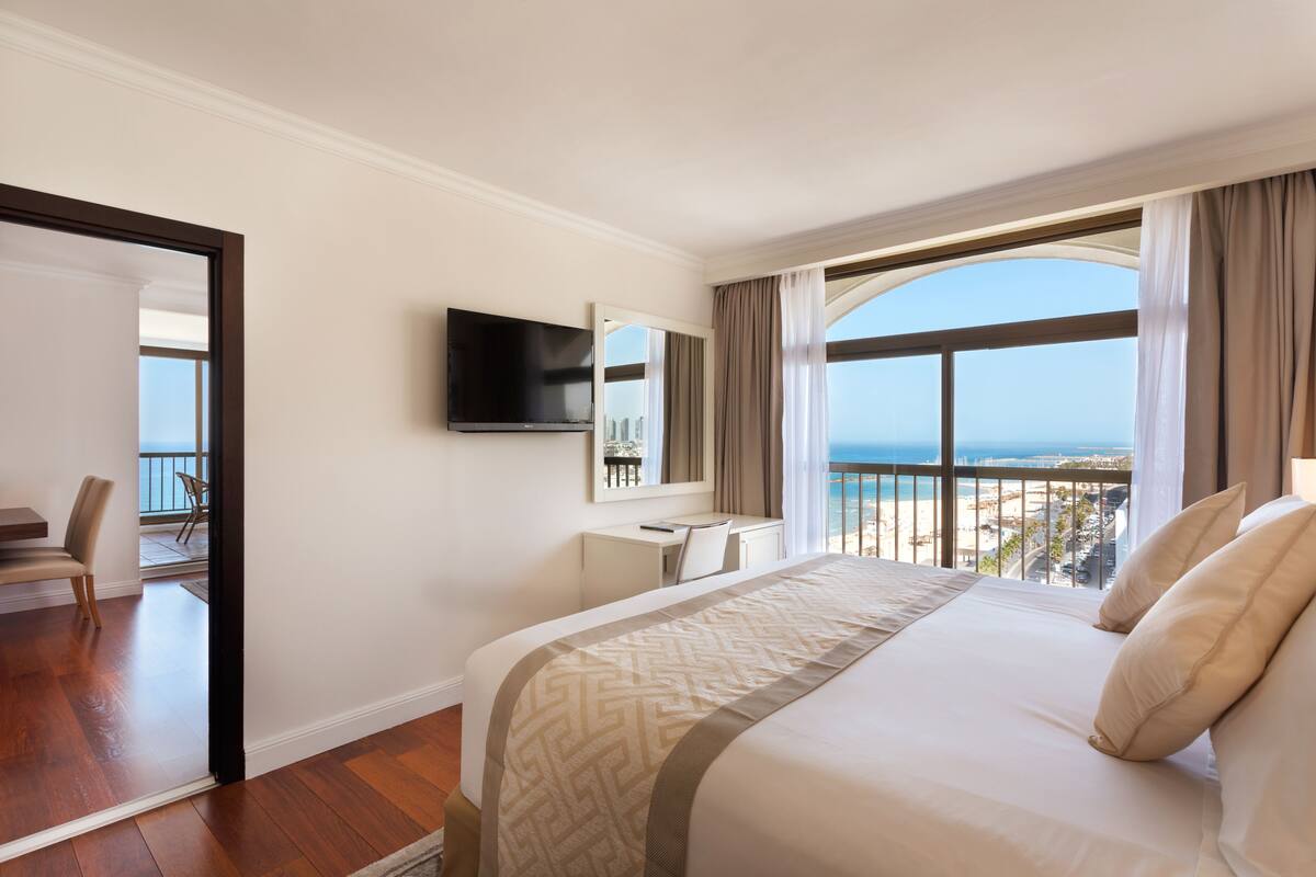 A hotel room with a large bed, a desk, a TV, and a balcony with a view of the sea.
