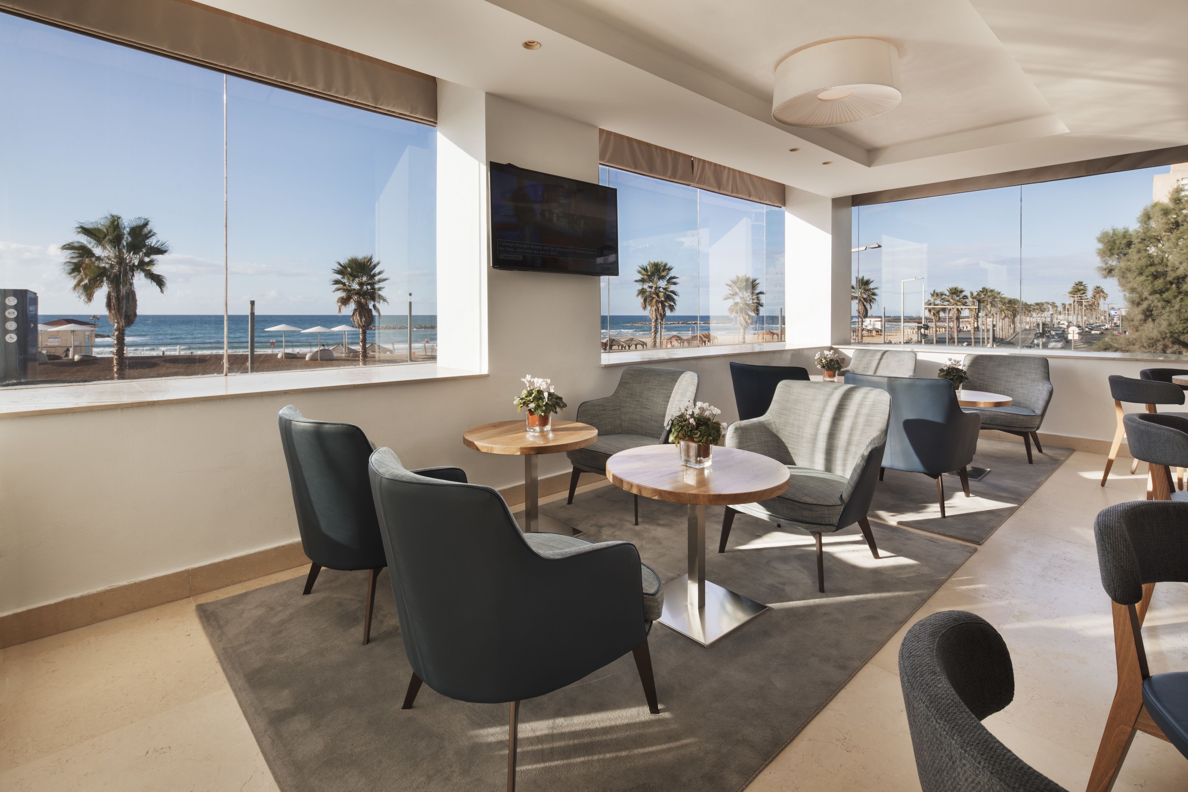 A modern hotel room with large windows offering a beach view, comfortable seating, and a flat-screen TV.