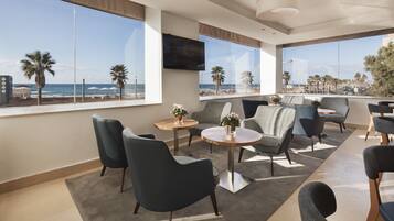 A modern hotel room with large windows offering a beach view, comfortable seating, and a flat-screen TV.