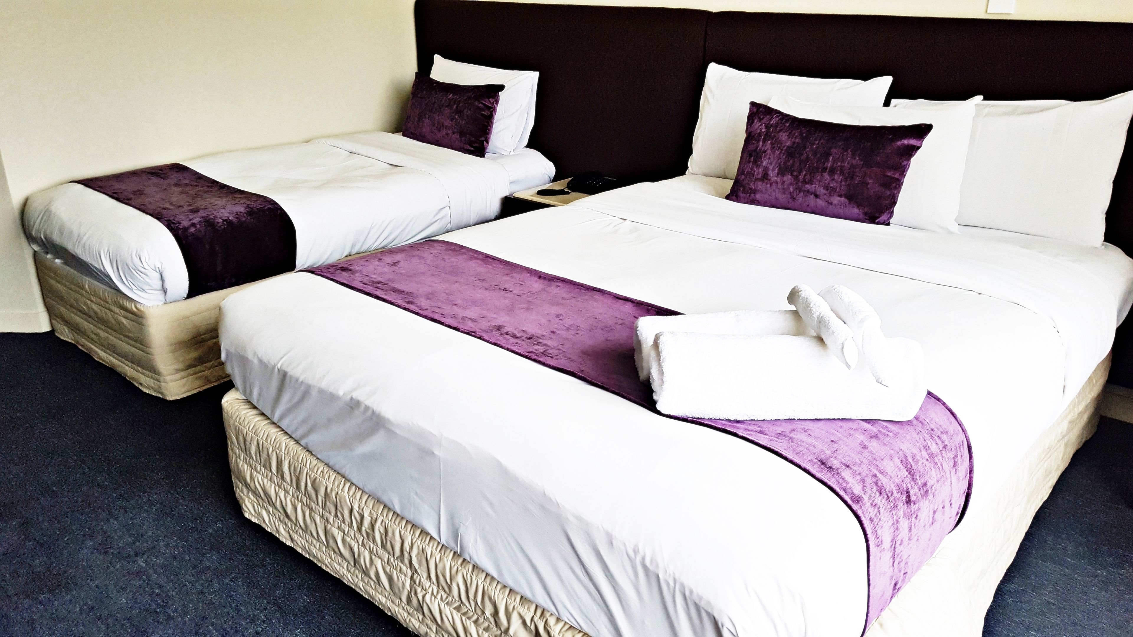 Two double beds with white and purple bedding, a dark headboard, and a small bedside table with a phone.